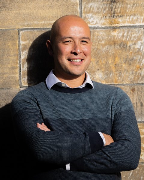 Akira O'Connor, smiling, arms folded, against a sandstone wall